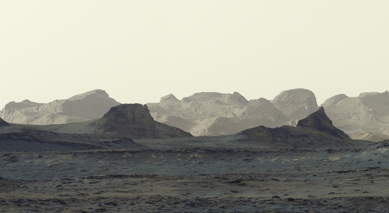 A stark, grayish-tan Martian landscape features jagged rock formations and rolling mountain ridges beneath a pale, hazy sky