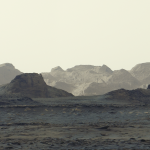 A stark, grayish-tan Martian landscape features jagged rock formations and rolling mountain ridges beneath a pale, hazy sky