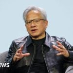 Jensen Huang, the CEO of Nvidia, is seated in a grey armchair against a plain, light-blue background